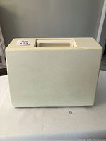 White hard plastic carrying case for the sewing machine with a molded handle on top. Case shows slight wear and aging.