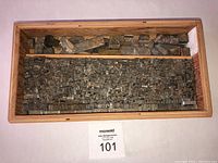 Top view showing metal typeset pieces densely packed in the larger compartment of the plywood tray, with larger metal blocks in the smaller compartment.