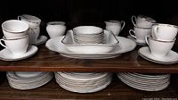 Full display of vintage white porcelain dinnerware with double gold line trim, arranged on wood shelf showing stacks of plates, cups and serving dishes.
