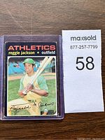 Card showing Reggie Jackson batting stance in Athletics uniform, encased in protective plastic sleeve, includes lot tag with number 58.