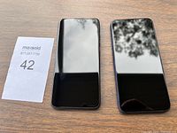 Two black touchscreen cell phones placed side by side on a wooden surface, showing full front panels with reflective glass screens.