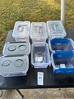 Top-down view of seven plastic storage containers arranged on a black table outdoors, showing various lid styles and colored handles.