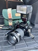 Canon EOS ELAN camera with attached Canon zoom lens, Sunpak flash on top, and green Canon carrying bag in background.