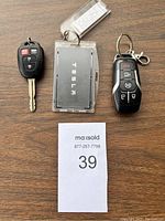 Top view of Tesla key card in transparent holder, Toyota key, and Ford key fob placed side by side on a wooden surface with a MaxSold lot number card.