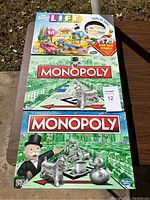 Three boxed board games placed outdoors on a table: The Game of Life on top, green edition Monopoly in the middle, blue edition Monopoly on bottom.