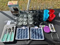 Photo showing large quantity of glassware including clear champagne flutes, clear drinking glasses, black tinted glasses, reusable cups, kitchen utensils in white tray, pot holders, silicone scrubber and tongs.