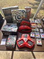Full overview photo of all games and accessories in the lot placed on a table outside, showing board games, poker chips, multiple decks of playing cards including Bicycle and UNO, and electronic UNO card accessories.