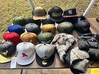 Large assortment of hats laid out on a table, showing various colors, brands and logos including Call of Duty, Hustle, New York Yankees, and others, alongside a fur-trimmed winter hat and gloves.