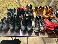12 pairs of men's shoes and boots spread on a table outdoors in natural light. Includes hiking boots, work boots, athletic sneakers, and slide sandals.
