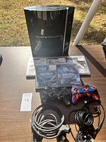 Photo showing Sony PlayStation 3 console, two controllers, and a stack of six PlayStation 3 game cases on a table outside in sunlight.