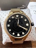 Close up front view of Puma gold tone wristwatch with black dial and mesh band on white pillow.