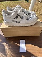 Side view of both white Nike Air Force 1 shoes showing blue Dior-inspired monogram pattern and HANNA embroidery on heel, placed on a cardboard box outdoors.