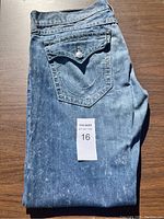 Pair of folded True Religion brand men's jeans on wooden surface, showing iconic back flap pocket with metal button and signature curved stitching, blue denim with light faded spots.