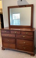 Front view of the dresser with attached mirror, showing the 9 drawers and brass handles in medium-dark wood finish.