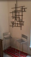 Pair of silver metal bar stools with horizontal bar backrests and footrests seen against a corner wall with the black abstract geometric metal wall sculpture mounted above and a red striped rug beneath the stools