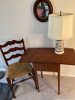 View of drop leaf table, wooden chair with thatched seat, blue and white floral lamp, and small round mirror on wall