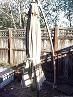 Closed beige fabric porch umbrella with brown frame, showing moss stains, secured with a strap, standing on an outdoor deck near wooden fence.