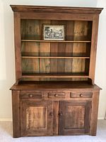 Full front view of the farmhouse wood hutch showing its shelves, drawers, and cabinet doors with vintage art placed on the top shelf.