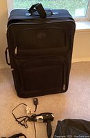 Large black soft shell rolling suitcase standing upright with front zippered compartments, hair dryer with cord on floor in front