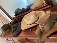 Multiple men's hats displayed on wooden dresser and reflected in mirror, showing variety of styles and materials