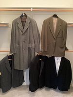 Wide shot showing four men's dress outerwear pieces hung on a rod and displayed in a closet space. Neutral shades including gray, taupe, camel, and black are visible. Jackets appear clean and lightly used.