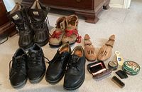 Four pairs of men's shoes including black dress shoes, tan boots, and black winter boots, with shoe care items and wooden shoe stretchers arranged around them on floor.