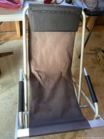 Brown fabric sling-back camp chair with metal frame, viewed folded and resting on the ground.