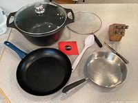 Photo showing all items: large black Fundix pot with glass lid, Bialetti frying pan with blue handle, Revere stainless steel pan, white serving spoon, set of steak knives in wooden block.
