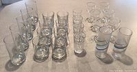 Overall photo of various glassware including short glasses, shot glasses, stemmed glasses and two tall textured hand blown glasses with colorful rainbow swirls.