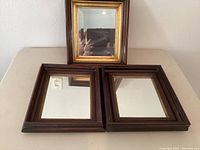 Three small wall mirrors resting together, two with plain dark brown wooden frames and one with a dark wooden frame and gold inner border.
