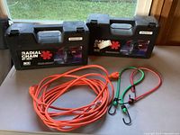 Two hard plastic cases labeled 'RADIAL CHAIN by SC' with coiled orange extension cord and two bungee cords on table.