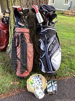 Two nylon golf bags standing against a tree outdoors, clubs visible inside the bags. A plastic bag full of golf balls in front and some packaged golf gloves on ground.