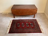 Vintage wooden coffee table with drop leaves folded down alongside a small decorative rug on a carpeted floor.