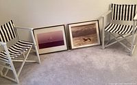 Two vintage white wooden beach chairs with blue and white striped fabric seats and backs next to two framed ocean photographs on carpet