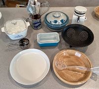 Overview of entire kitchenware lot showing all items on countertop including Pyrex, Corning Ware, pottery pie pan, colander, measuring cups, utensil holder with whisk and other utensils