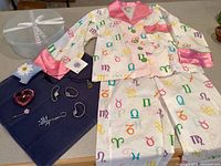 Full view of new women's pajama set with zodiac print, satin pink collar and cuffs, displayed alongside several women's accessories: watches, necklaces, bracelets, and keychain on a dark blue cloth next to a round box.