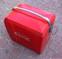 Red rectangular metal vintage Coca-Cola cooler showing exterior with metal handle and embossed logo.