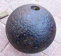 View of antique iron cannon ball showing textured, rusted surface and recessed mark on one side.