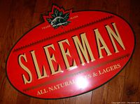 Close-up image of the front of a large oval Sleeman beer metal sign with red background, gold lettering, and a green maple leaf and beaver trademark.