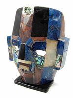 Front angled view showing the colorful stone inlay and geometric facial features with cutouts for eyes and mouth, mounted on black base.