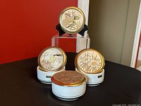 Photo of four round porcelain trinket dishes with white bases and lids decorated with gold accented Japanese Chokin style bird and flower designs arranged on clear and white stands against a red background.