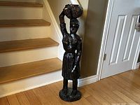 Full view of large hand-carved wooden female statue with basket on head next to staircase and door.