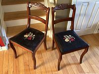 Photo showing pair of antique wooden chairs with carved ladder backs and embroidered floral seats against a hardwood floor.