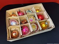 All 12 glass Christmas ornaments displayed in the original divided box, showing a range of colors and designs including pink, gold, red, orange, silver, and some with glittery floral painted details.