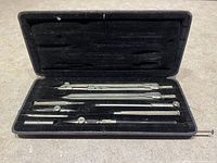 Open black hard case with seven metal drafting tools arranged inside, set on concrete surface.