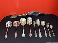 Top view of 11 antique silver spoons and serving utensils laid out on black surface with red background
