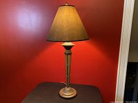 Front angle view of bronze metal table lamp with dark brown conical fabric shade, showing base, column with studs, and shade detail.