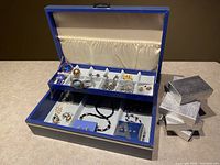 Full view of blue jewelry box filled with assorted jewelry and several silver textured jewelry boxes beside it.