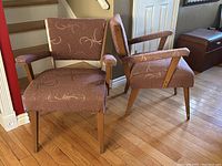 Pair of mid century modern armchairs with dark rose fabric upholstery featuring swirled zig zag pattern, wooden legs and armrests, placed on hardwood floor against a wall and stairs.