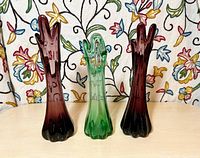 Front view of three swung glass vases, two purple and one green, on a table with a floral patterned fabric background.